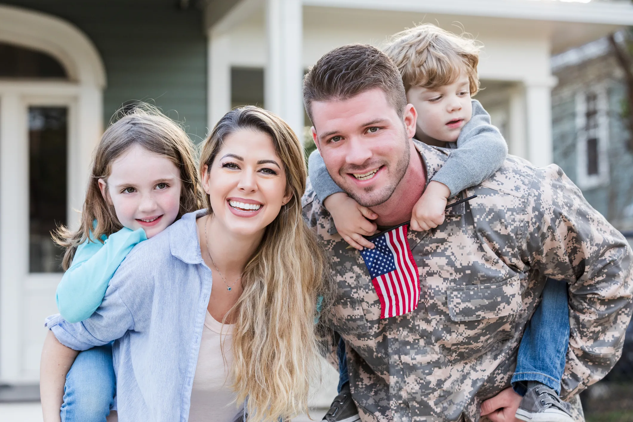 A smiling Army service member and his wife outside their home, each carrying one of their two children piggyback, a small American flag in hand