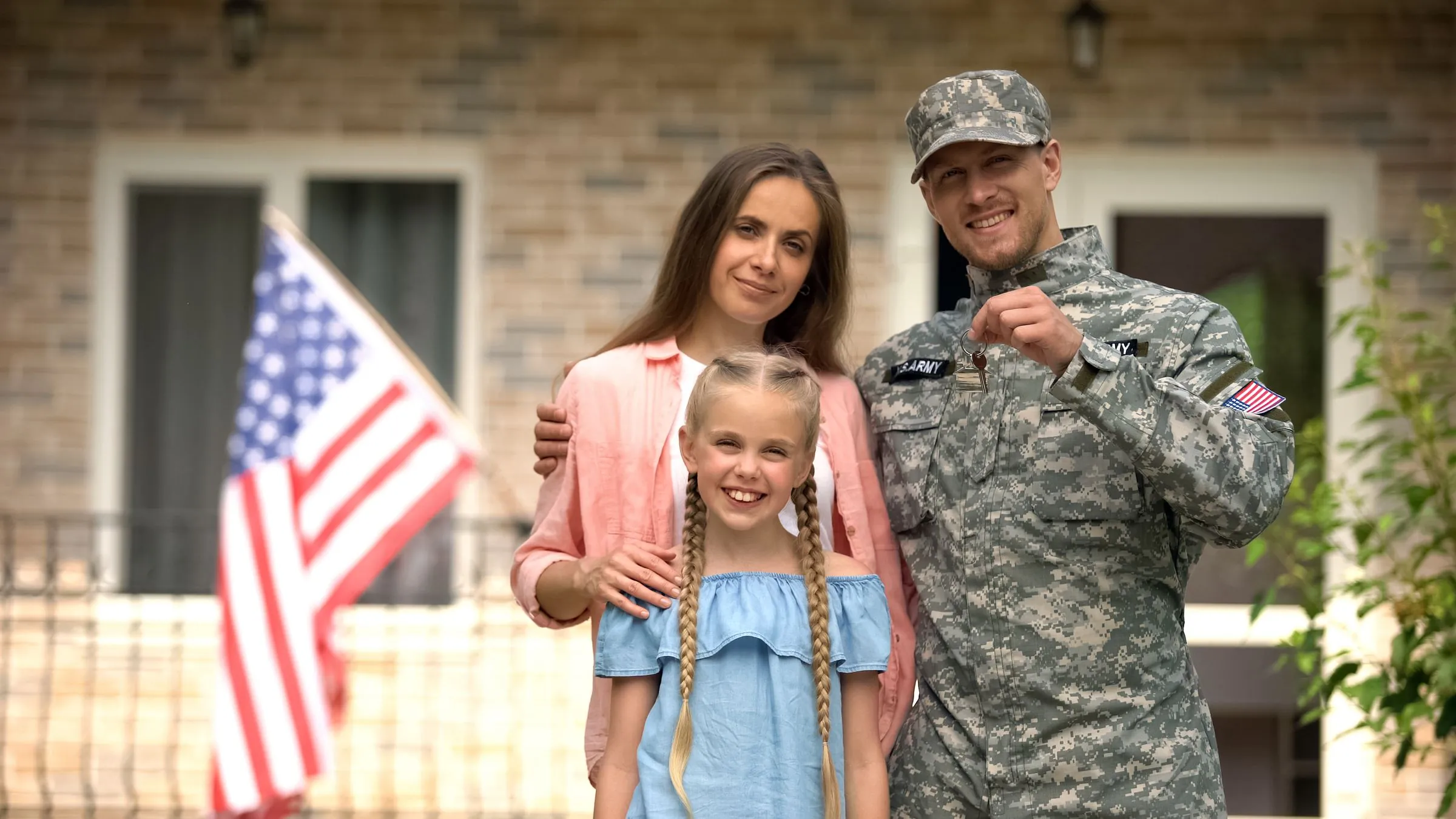 An Army service member with his wife and daughter outside their home, an American flag behind them