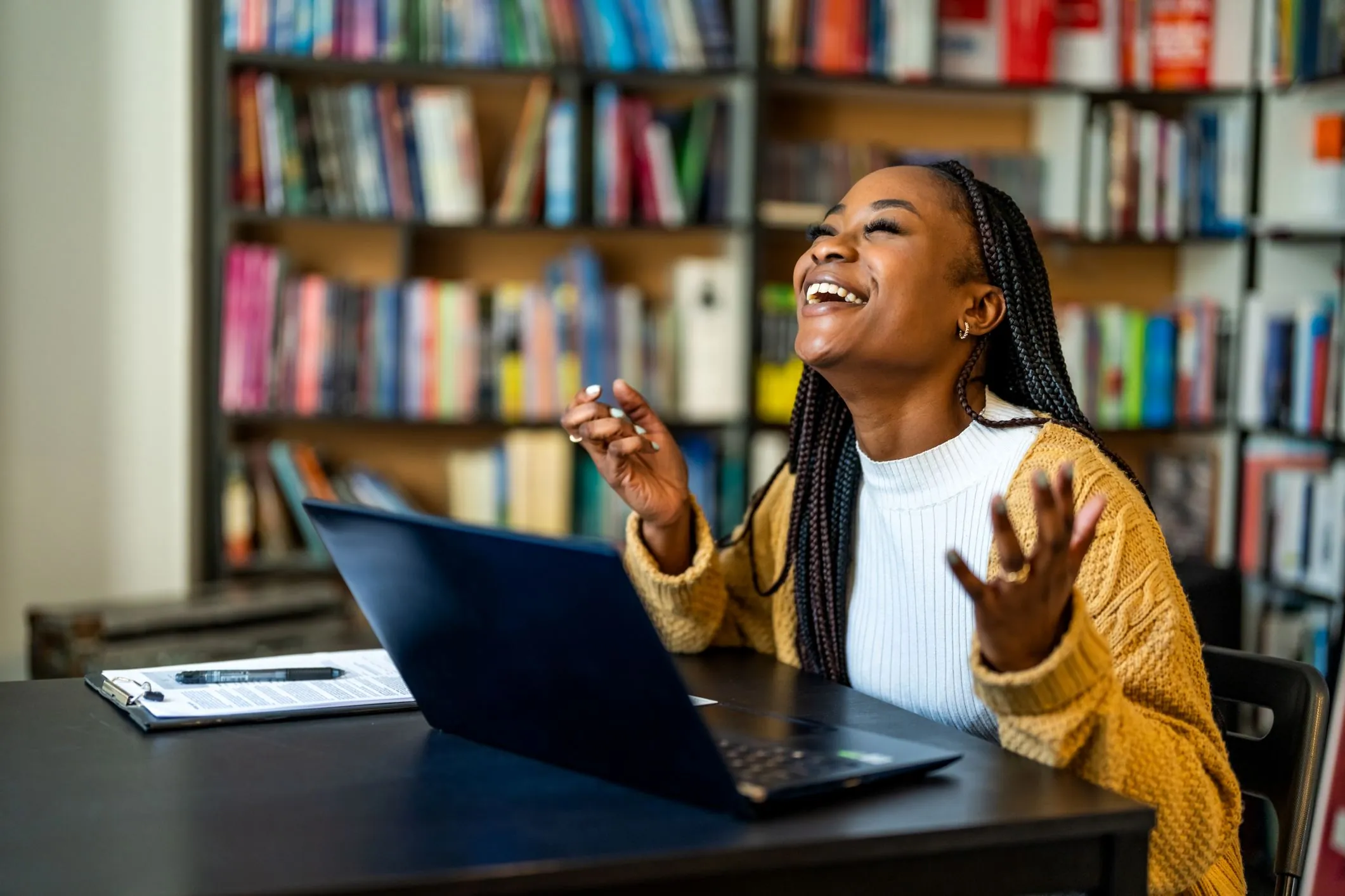 A student laughing with her arms up at a laptop in front of library bookshelves